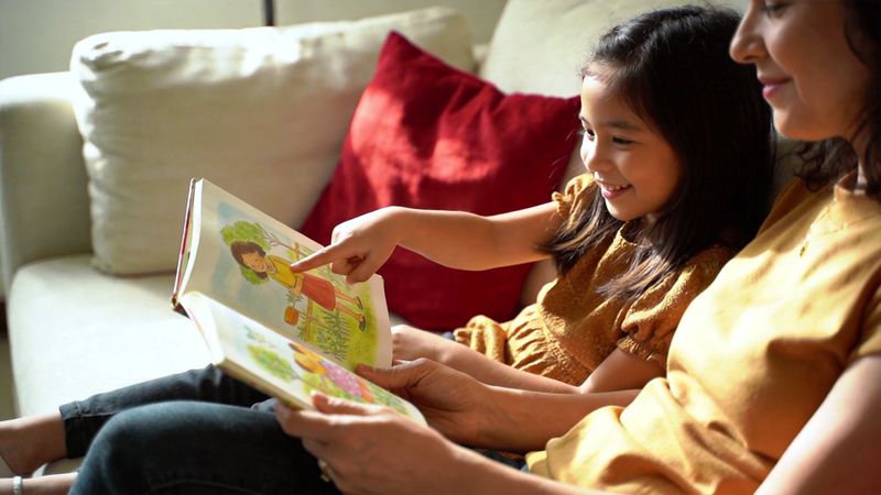 Mother and daughter reading a picture book together on a sofa, the girl pointing at an illustration